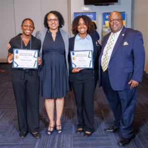 Four people stand together smiling; two women in business attire hold certificates. Another woman and a man in formal wear stand beside them in an indoor setting, likely at an award or recognition event.