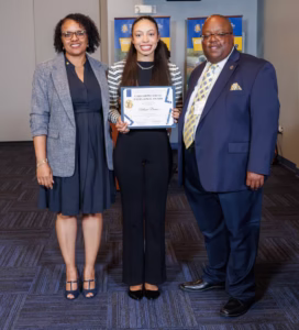 Three people stand indoors, smiling at the camera. The woman in the center holds a certificate. Two others, a woman on the left and a man on the right in a suit, stand beside her. Blue and yellow banners are in the background.