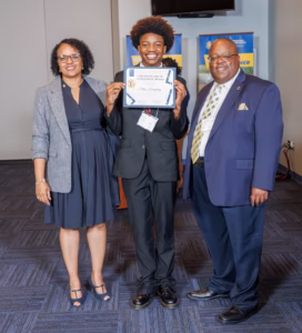 Three people stand indoors; the person in the center, smiling, holds up a certificate. The two on either side are also smiling. All are dressed in business attire, with blue and gray tones in the background.