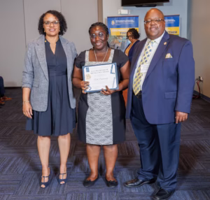 Three adults in business attire stand together indoors. The woman in the center holds a certificate and smiles at the camera. Two banners with text and images are visible in the background.