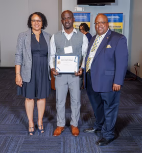 Three people stand indoors, smiling at the camera. The man in the middle holds a certificate. The two people on either side are dressed in business attire, and posters are visible in the background.