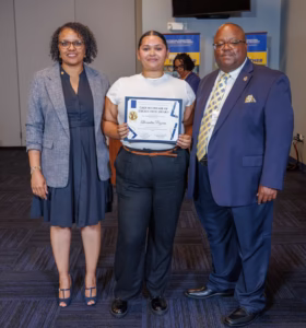 Three people stand indoors, smiling at the camera. The person in the center holds an award certificate. The two others, a woman and a man in business attire, stand on either side of the recipient.