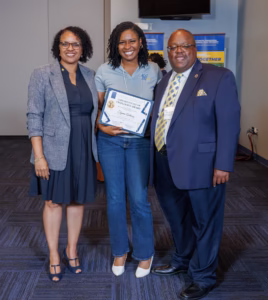 Three people stand indoors, smiling at the camera. The woman in the middle holds a certificate. All are dressed in business or business-casual attire, with banners and a TV screen visible in the background.
