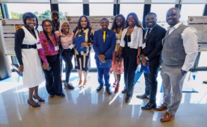 A group of ten people dressed in business or professional attire stand smiling together indoors, with research posters displayed behind them and sunlight streaming through large windows.
