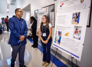 A man and a woman stand and smile near a large research poster in a hallway, with other people and posters visible in the background at what appears to be an academic event or conference.