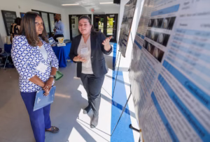 A woman in a blazer gestures while explaining a research poster to another woman holding a folder at an indoor event. Other people and display tables are visible in the background.
