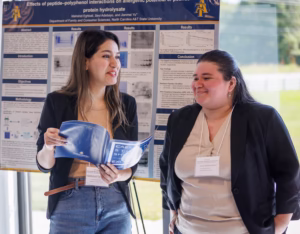 Two women stand in front of a scientific poster, smiling and talking. One woman holds a booklet and gestures as she speaks, while the other listens with hands in her pockets. Both wear name badges and casual business attire.