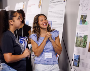 Two young women stand in front of research posters at an event. One smiles and gestures while presenting, wearing a name tag and holding her hands together. The other listens, holding a folder and papers.