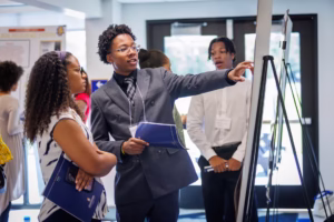 A young man in a suit points at a display board while talking to a woman holding a booklet. Other people stand nearby in a well-lit room with posters and large windows in the background.