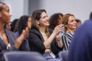 A woman in a black blazer smiles and claps while sitting among a group of people at an indoor event, with others around her also applauding and appearing engaged.