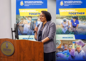 A woman speaks at a podium with a microphone in front of banners for North Carolina Agricultural and Technical State University’s Agriculture and Environmental Sciences, featuring the slogan Growing... Together.