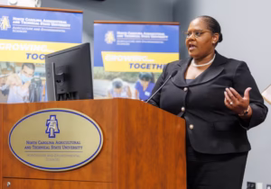 A woman in professional attire speaks at a podium with the North Carolina Agricultural and Technical State University logo. Banners behind her display the schools name and Growing... Together.