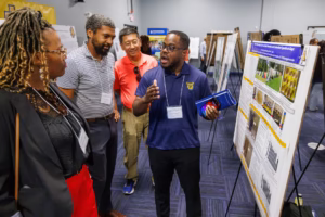 A group of four adults stands by a research poster at an indoor event. One man gestures while explaining the poster; the others listen attentively. Poster boards and other attendees are visible in the background.
