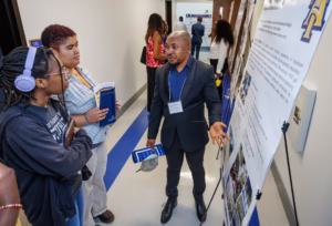 A man in a suit explains a large poster to two attentive students in a hallway during a school event. Other people are seen walking and conversing in the background.