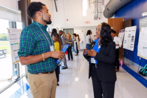 Two people have a conversation in front of research posters at a bright indoor academic event, while others interact and view posters in the background.