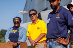 Three people stand outdoors in sunlight. One man in a yellow shirt and sunglasses smiles at the camera, while two others in blue shirts stand beside him. A modern building and blue sky are visible in the background.