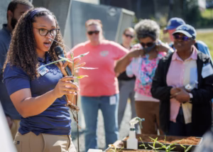 A woman with curly hair and glasses holds a plant and speaks into a microphone while demonstrating to a group of attentive adults outdoors. A tray of seedlings and a spray bottle are on the table in front of her.
