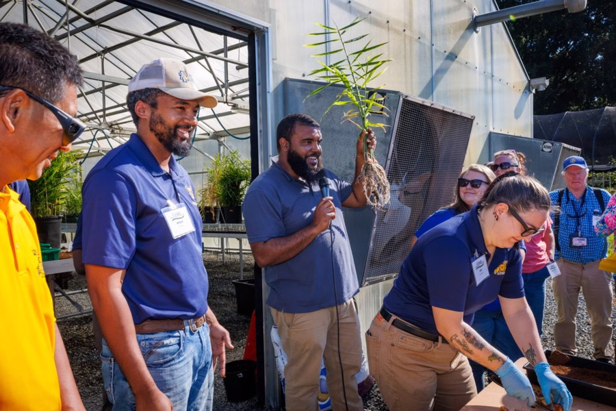 A group of people gather outside a greenhouse. One person holds up a large plant root and speaks, while others in blue shirts observe or work with plants on a table. It’s sunny and everyone looks engaged.