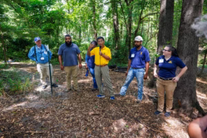Six people stand in a wooded outdoor area, some wearing name tags and work uniforms. One person in a yellow shirt speaks into a microphone while others listen attentively. Sunlight filters through the trees above.