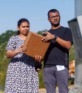 Two people stand outdoors. The woman in a floral dress holds a large, brown, textured cube, while the man in glasses and a black shirt gestures and speaks, appearing to explain something about the object.