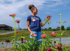 A woman wearing a blue t-shirt and jeans stands outside smiling, holding a pink flower among blooming flowers, with a grassy field, buildings, and a blue sky in the background.