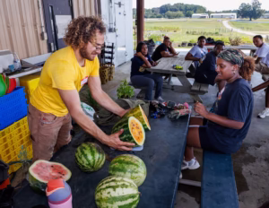 A person in a yellow shirt shows a sliced watermelon with yellow flesh to a group of people seated at a picnic table outdoors; other watermelons and supplies are on the table.