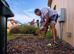 A group of people are working together outside by a beige building, pulling weeds and tending to a garden. One person in the foreground is bending over, wearing gloves, athletic clothes, and yellow sneakers.
