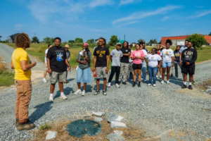 A group of people stands outdoors on a gravel path, listening to a person in a yellow shirt speaking. The group appears engaged, with some holding notebooks. The sky is clear and grassy fields surround them.
