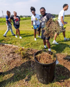 Five people participate in a community clean-up; one man dumps a pile of weeds into a black trash bin while others stand nearby on a grassy field under a clear sky.
