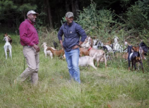 Two men stand in a grassy field, smiling and talking, surrounded by a herd of goats grazing near the edge of a wooded area.