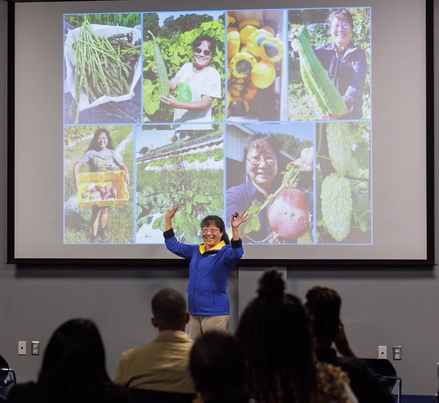 09_17_2025_ABIC 001 A woman speaks enthusiastically to an audience in front of a screen displaying a collage of photos showing her harvesting and holding various vegetables on a farm.