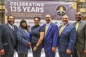Six professionally dressed people stand together smiling in front of a banner that reads “Celebrating 135 Years” with a logo for the 1890 Land-Grant Universities and their anniversary.