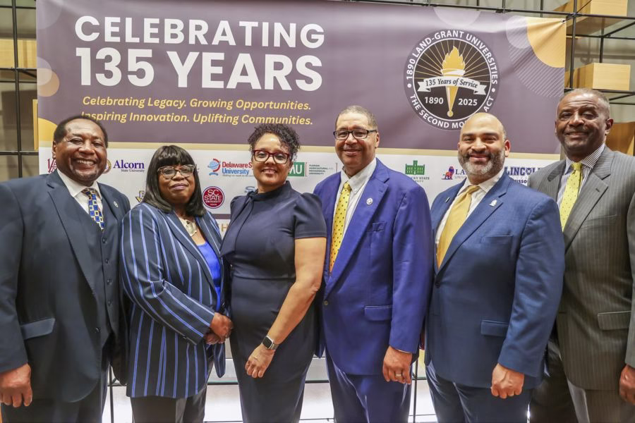 Six professionally dressed people stand together smiling in front of a banner that reads “Celebrating 135 Years” with a logo for the 1890 Land-Grant Universities and their anniversary.