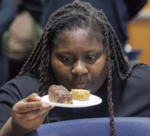 A person with long, braided hair examines two desserts on a plate, looking contemplative. The background is blurred, focusing on the individual and the treats.