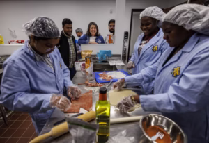 A group of people in lab coats prepare food in a kitchen. Two participants are working on dough, while others observe and assist, creating a collaborative cooking environment.