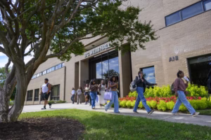 Students walk along a sidewalk in front of B.C. Webb Hall, a tan brick building with large windows, on a sunny day. Trees and colorful flowers are visible beside the walkway.