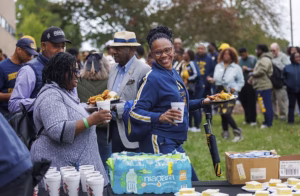 A group of people at an outdoor gathering, smiling as they hold plates of food and drinks. Tables with cups and snacks are visible in the foreground, and others are mingling in the background.