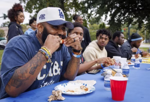 A group of people sit at a long table outdoors, enjoying food together. The man in the foreground, wearing a blue LA cap and bracelets, eats from a plate while others chat and eat beside him. Cups and water bottles are on the table.