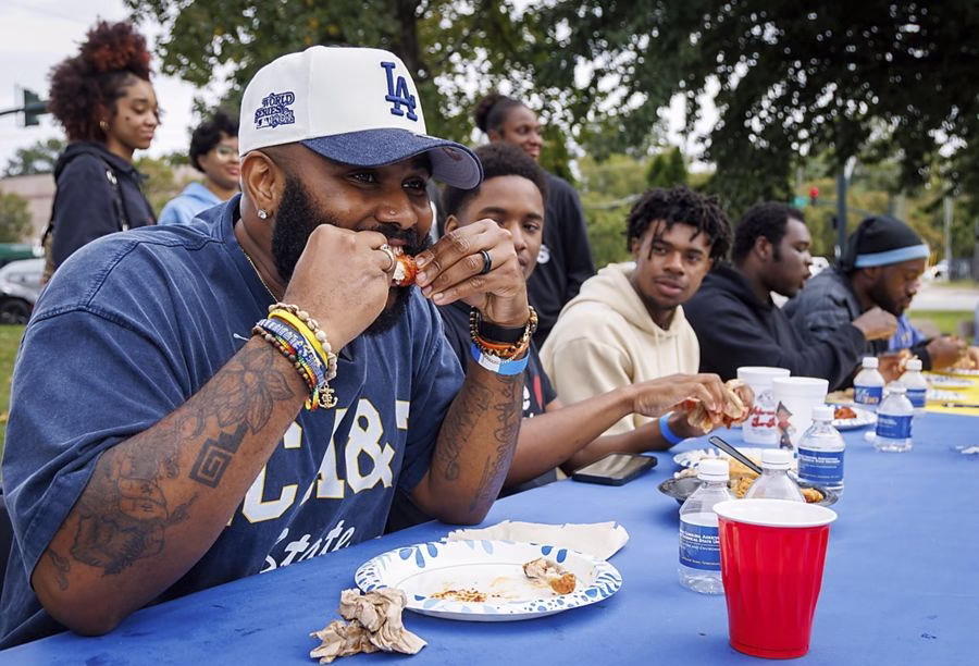 A group of people sit at a long table outdoors, enjoying food together. The man in the foreground, wearing a blue LA cap and bracelets, eats from a plate while others chat and eat beside him. Cups and water bottles are on the table.