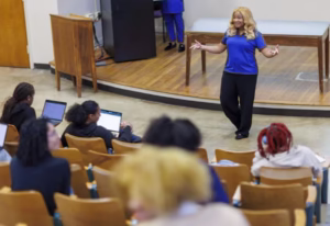 A woman with long, wavy hair speaks engagingly to an audience in a lecture hall. Students are seated, some using laptops, while she gestures animatedly. A table is positioned behind her.
