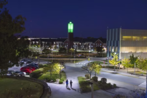 A well-lit campus scene at night featuring two people walking on a path. In the background, a tall green-lit tower stands near a modern building, with parked cars and trees in the surroundings.