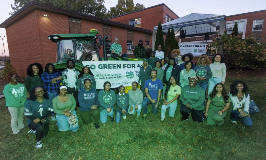 A diverse group of people in green attire gathers outdoors in front of a building, celebrating 4-H week with a tractor and a banner that says "Go Green for 4-H.