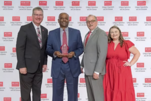 Four people stand smiling in front of an NC State College of Agriculture and Life Sciences step-and-repeat banner. The man second from the left holds a clear award. The group is dressed in business attire, with one woman in a red dress.