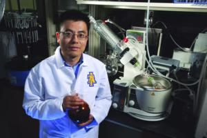 A scientist in a white lab coat stands in a laboratory, holding a round-bottom flask. Laboratory equipment and glassware are visible on the workbench behind him. The lab coat features a blue and gold A logo.