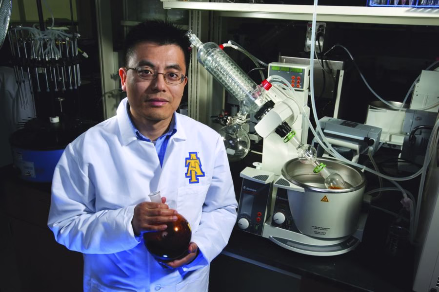 A scientist in a white lab coat stands in a laboratory, holding a round-bottom flask. Laboratory equipment and glassware are visible on the workbench behind him. The lab coat features a blue and gold A logo.