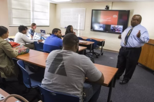A teacher in a dress shirt and tie speaks to students seated at desks in a classroom, with a presentation displayed on a screen behind him and notes written on a whiteboard.
