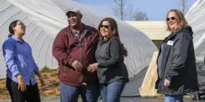 Four individuals smiling and interacting outdoors near a greenhouse, with one person gesturing while the others laugh. The scene conveys a sense of camaraderie and enjoyment.