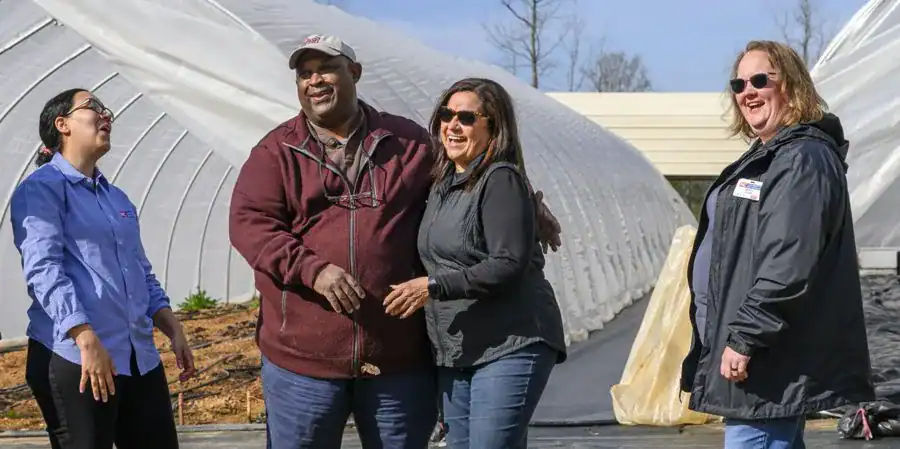 Four individuals smiling and interacting outdoors near a greenhouse, with one person gesturing while the others laugh. The scene conveys a sense of camaraderie and enjoyment.