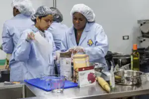 Two people in blue lab coats and gloves prepare food in a kitchen, focused on mixing ingredients. Various baking supplies and equipment are scattered on the counter.