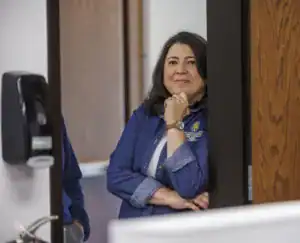 A woman with wavy hair leans against a doorway, smiling softly with her chin resting on her hand. She wears a denim shirt and a watch, and the background features a wood-paneled wall and a soap dispenser.
